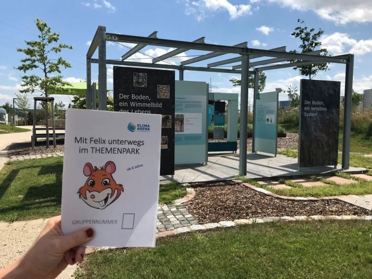 Eine Person h&auml;lt ein Natur-Rallye-Heft vor einem Outdoor-Infobereich im Themenpark hoch.