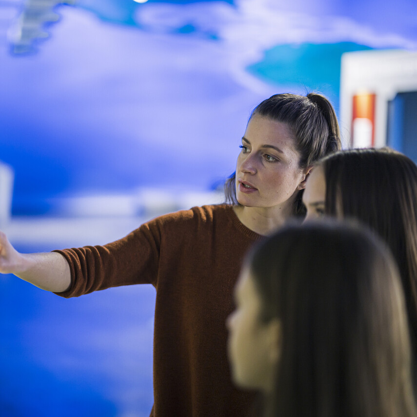 Lehrkraft in der Ausstellung mit Sch&uuml;lerinnen beim Bedienen eines Exponates.
